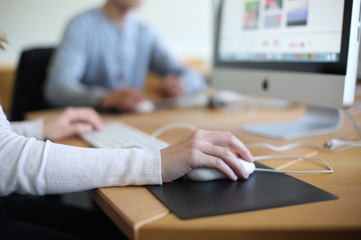 Die Nahaufnahme fokussiert auf eine Hand, die eine Computermaus auf einem dunklen Pad bedient, während im Hintergrund eine weitere Person unscharf an einem Monitor arbeitet. Die Szene verdeutlicht die Nutzung moderner IT-Arbeitsplätze in einer hellen Lernumgebung.