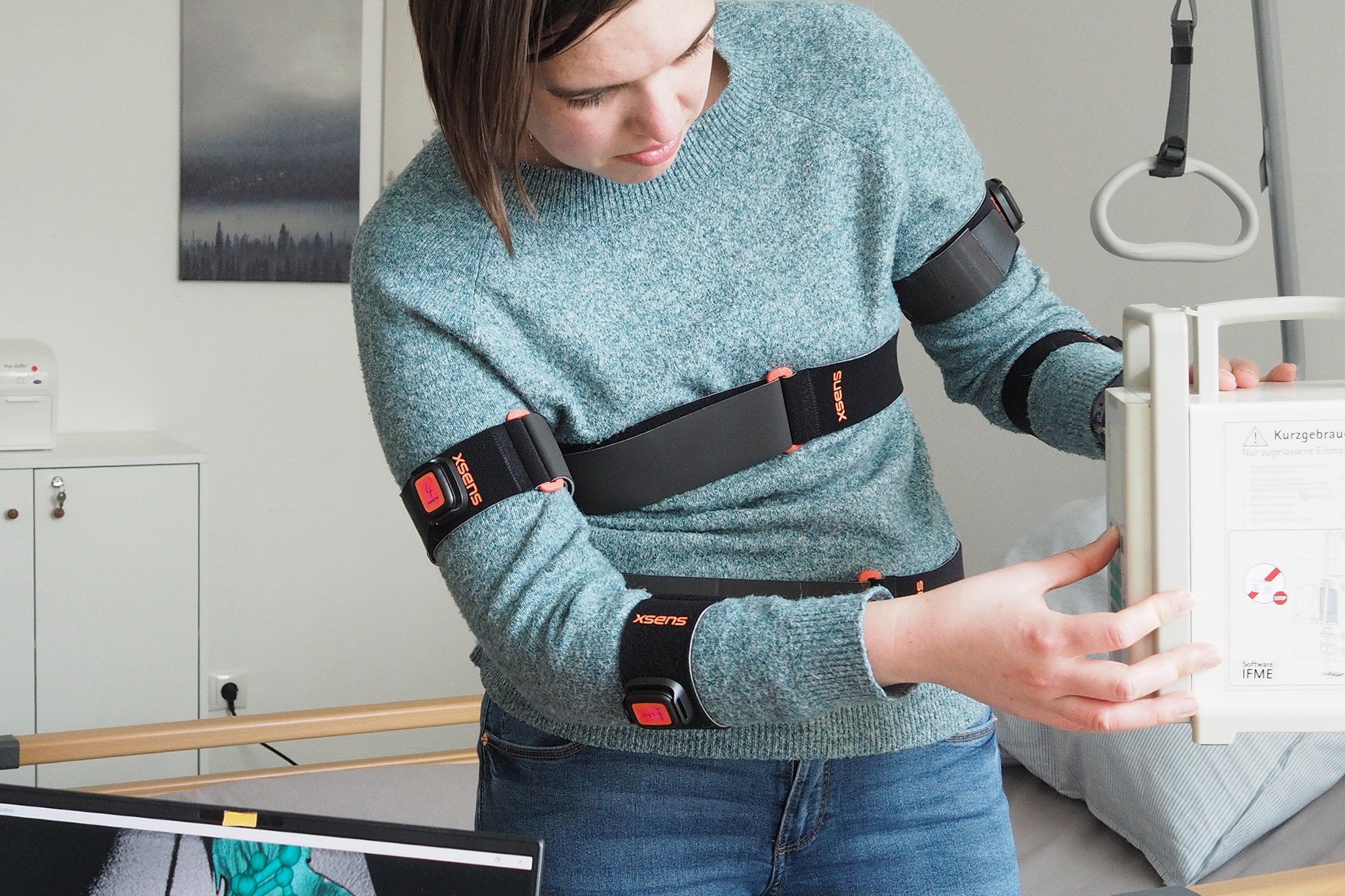 Eine Person ist mit Sensoren ausgestattet. Ihre Bewegungen werden mit einem Laptop aufgezeichnet. Im Hintergrund sieht man ein Krankenhausbett.