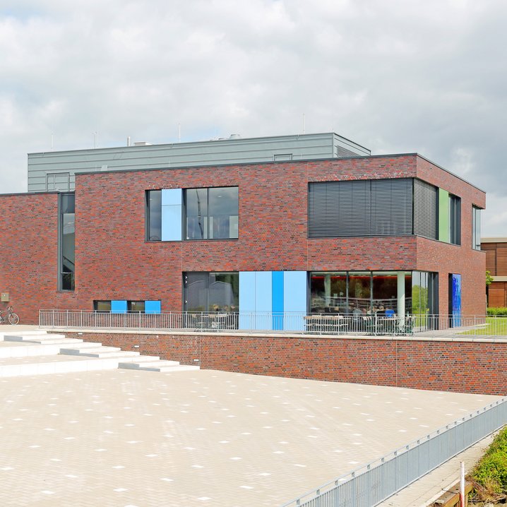 Das moderne Bibliotheksgebäude am Campus Elsfleth besticht durch eine rote Backsteinfassade, die durch bunte Paneele in Blau- und Grüntönen an den Fenstern aufgelockert wird. Im Vordergrund erstreckt sich ein großer, gepflasterter Platz mit flachen Stufen, der direkt zum Eingang des zweistöckigen Gebäudes führt.