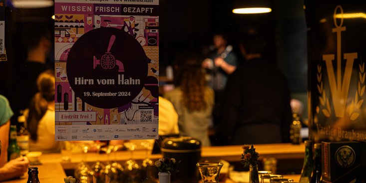 Im Vordergrund ist ein Plakat des Kneipenabends "Hirn vom Hahn" zu sehen. Dahinter sind der Kneipenraum und ein Vortragender.