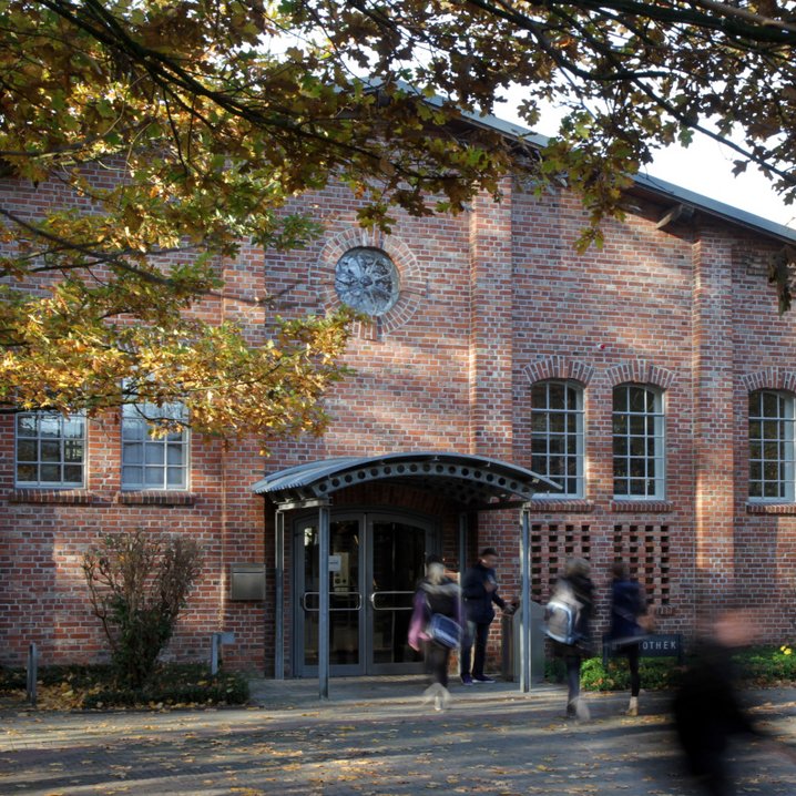 Die Aufnahme zeigt den Eingangsbereich einer Bibliothek in einem historischen Backsteingebäude mit Rundbogenfenstern und einem kleinen Vordach über der Tür. Im Vordergrund sind schemenhaft Passanten in Bewegung zu sehen, während herbstliche Bäume das Gebäude einrahmen.