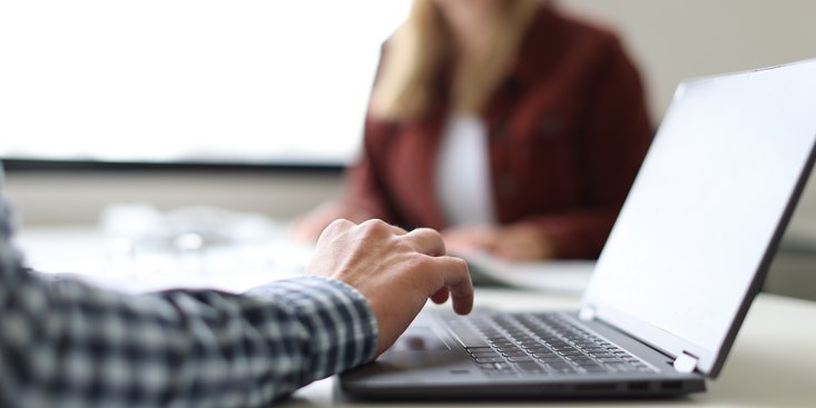 Eine Person tippt auf einem Laptop, der auf einem Tisch in einem hellen Raum steht. Im Hintergrund sitzt eine weitere unscharfe Person gegenüber, was auf ein Gesprächs- oder Arbeitsszenario hindeutet.