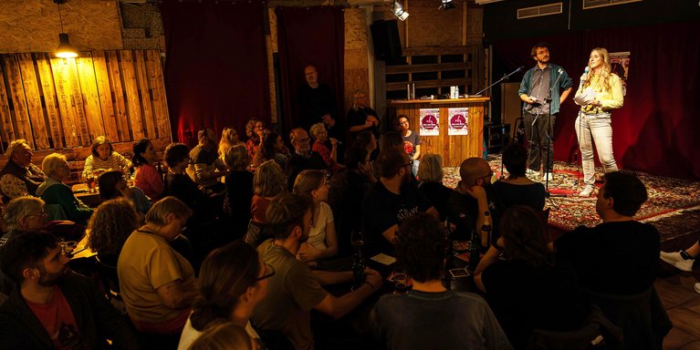 Zwei Personen stehen in einer Kneipe auf der Bühne. Vor ihnen sitzen viele Zuhörerinnen und Zuhörer.