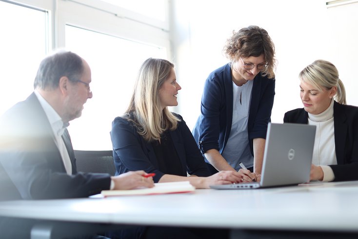 Eine Gruppe von vier Menschen in formeller Kleidung sitzt an einem Tisch um einen Laptop und arbeitet zusammen.